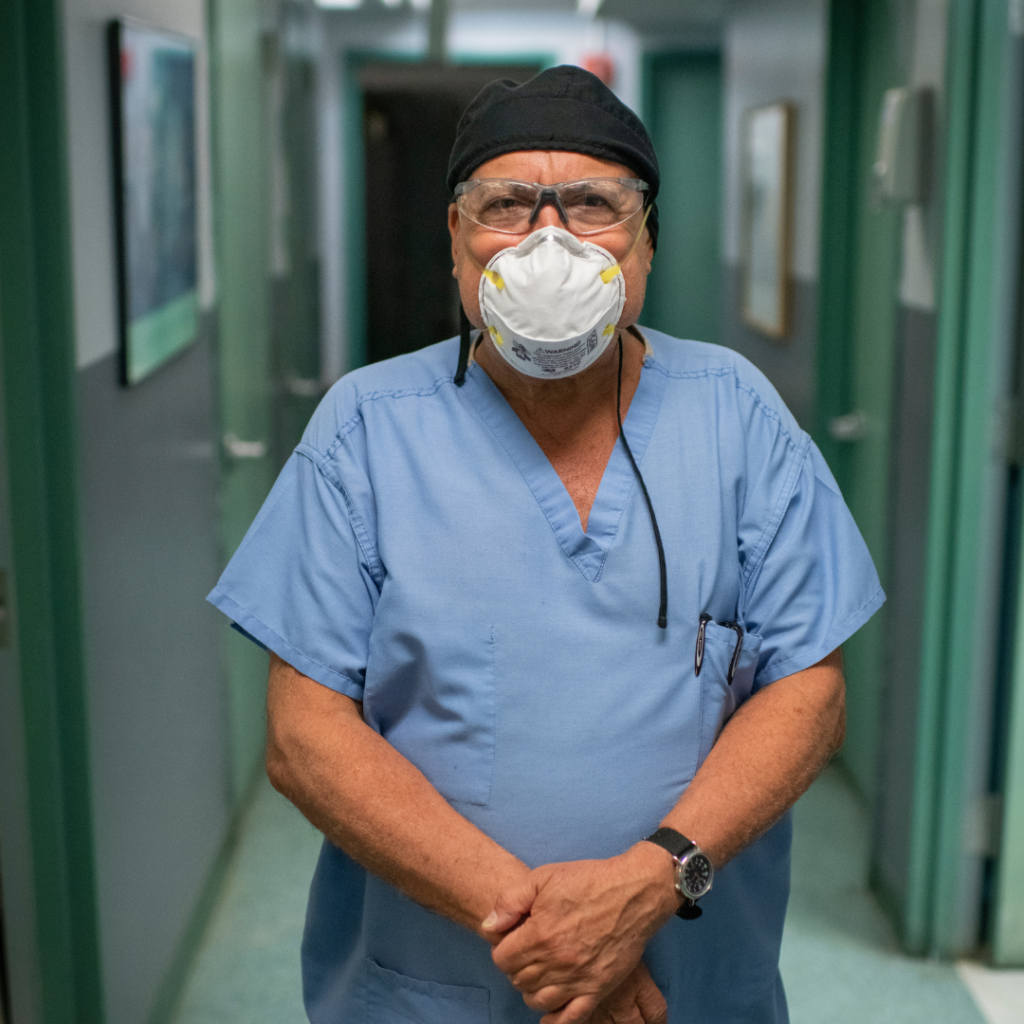Project Renewal’s longtime dentist, Dr. Mark Shufman, stands in the hallway of the Fort Washington Dental Clinic with his hands clasped in front of him. He is wearing an N95 mask and blue scrubs.