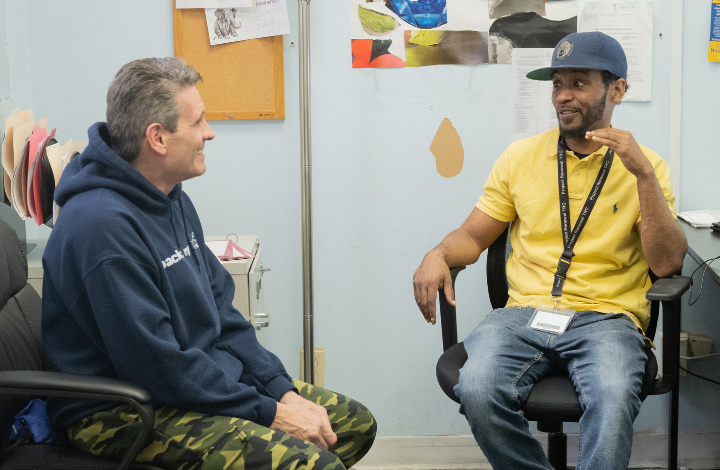 A staff member and a client in recovery sit across from each other talking and smiling inside a Project Renewal shelter.